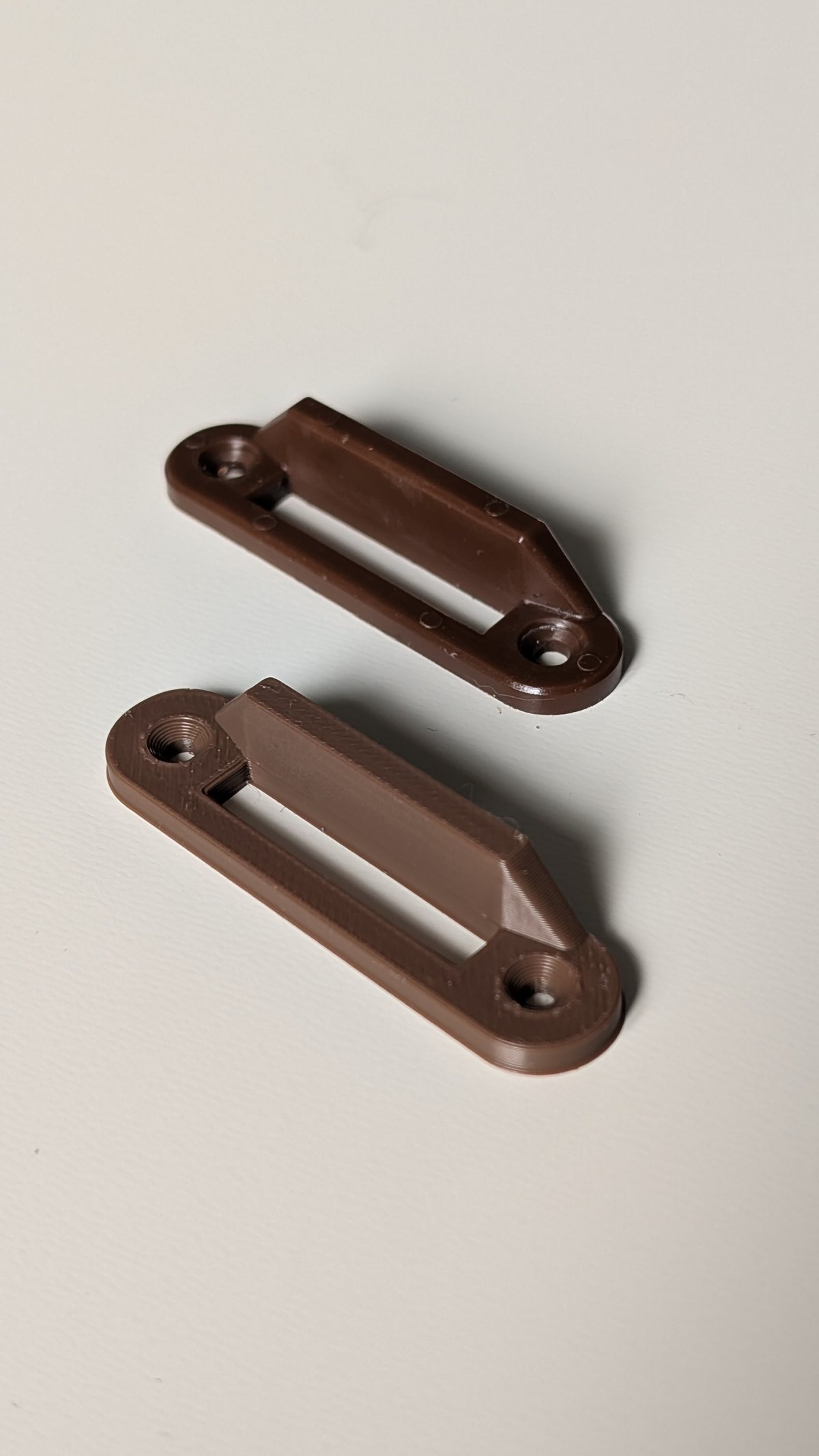 Deux verrous de meuble marron identiques posés côte à côte : l'un est la pièce d'origine moulée, l'autre la copie imprimée en 3D.
